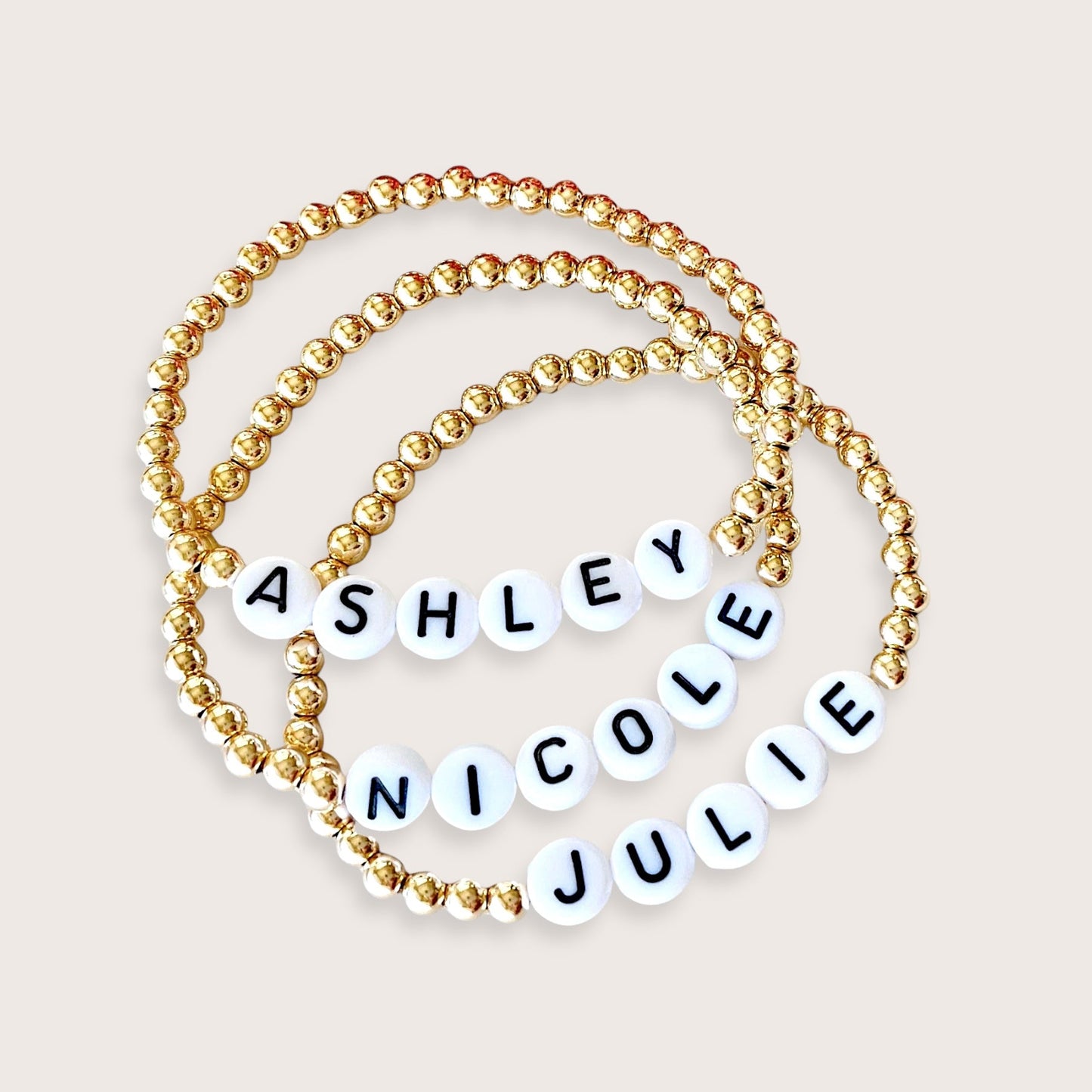 A gold-colored beaded bracelet with personalized engraved names 'Ashley Nicole' on it, showing a series of round beads strung on an elastic cord.