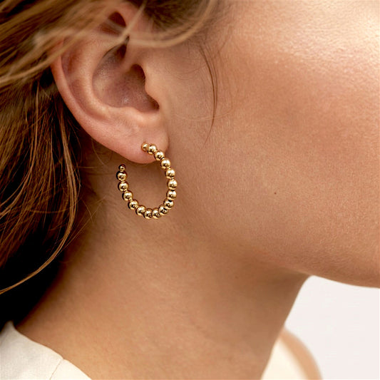 Close-up of a gold beaded C-shaped hoop earring worn in an earlobe, with brown hair tucked behind the ear.