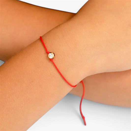 Red string bracelet worn on a wrist, showing the small gold bezel-set cubic zirconia between two red knots