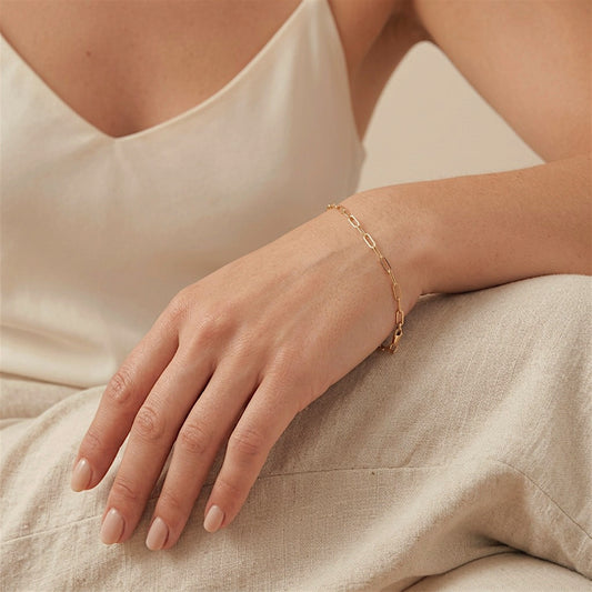 Delicate gold paperclip bracelet worn on a woman’s wrist, with her hand resting on ivory linen