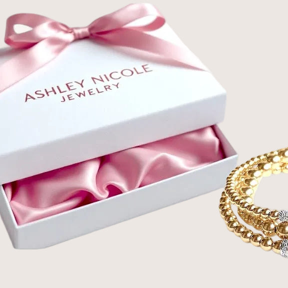 Gold bracelets on a white surface with an open jewelry box featuring a pink ribbon and 'Ashley Nicole Jewelry' branding.