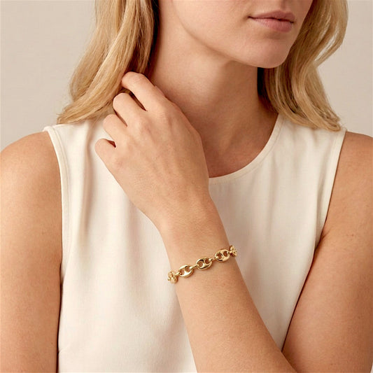 Blonde woman in an ivory sleeveless top wearing a gold puffed mariner bracelet, with her hand resting near her hair.