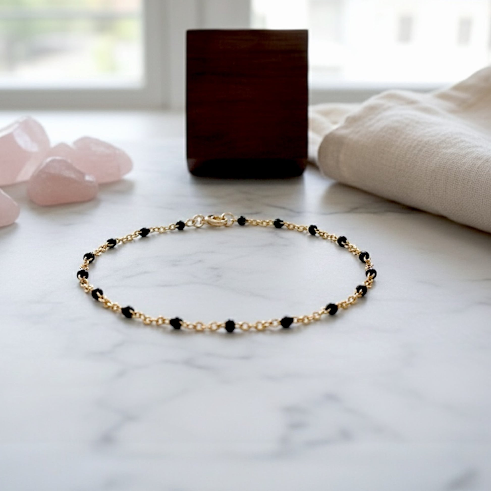 Gold bracelet with black beads on a marble surface with pink stones and a wooden block in the background.