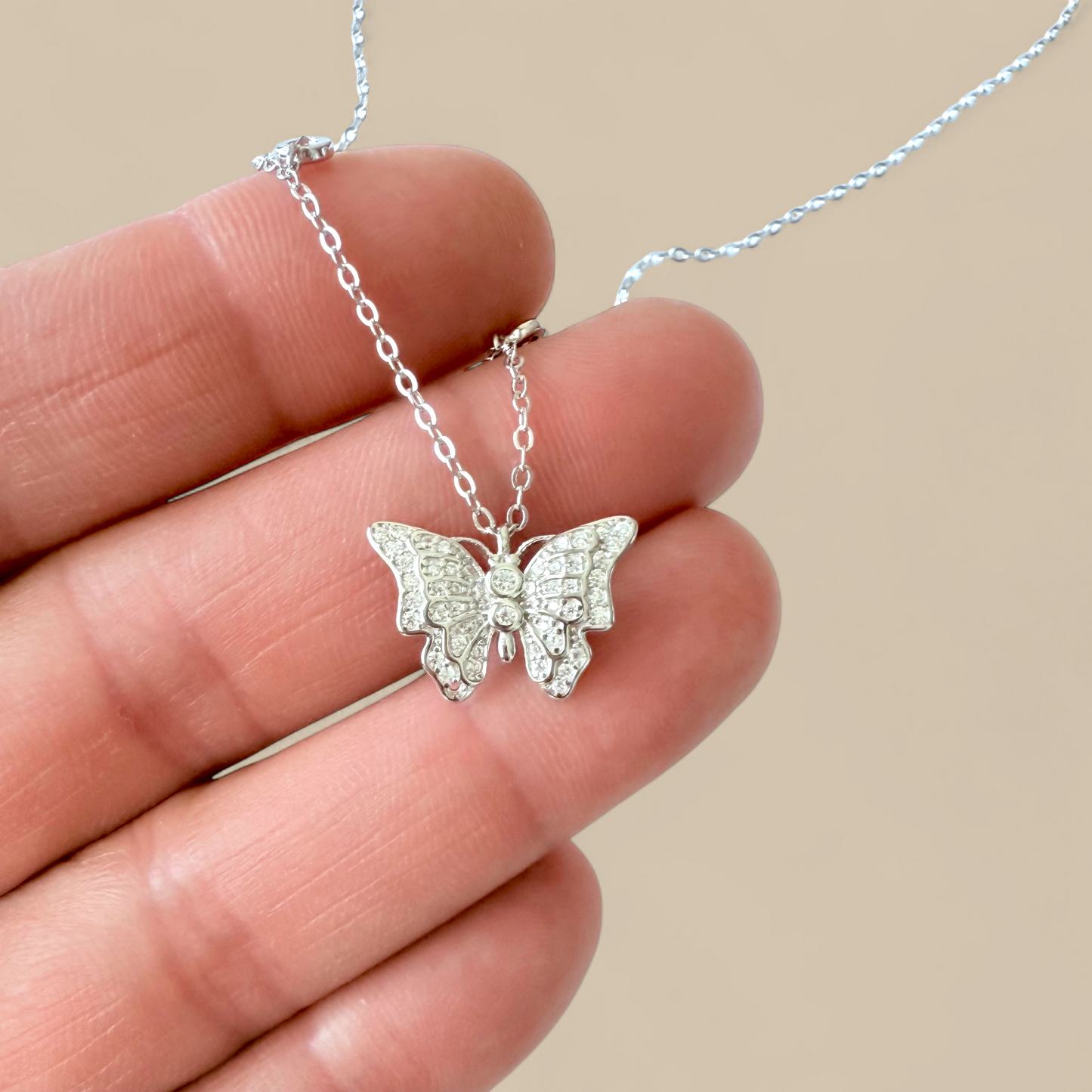 Silver butterfly necklace held between fingers against a beige background