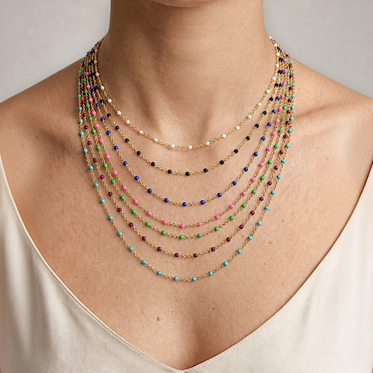 A model wearing a layered multi-strand beaded necklace. The necklace features multiple gold-tone chains with small colorful beads in blue, green, pink, purple, and black, along with a few white pearl-like beads.
