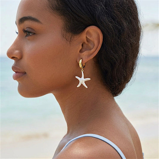 Woman wearing a gold huggie earring with a dangling white starfish charm, with the ocean in the background