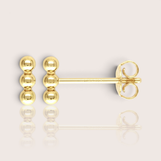 Close-up profile 14K gold-filled triple bead vertical bar stud earrings, display the minimalist, delicate gold cluster stud. The background is a soft, minimalist studio setting