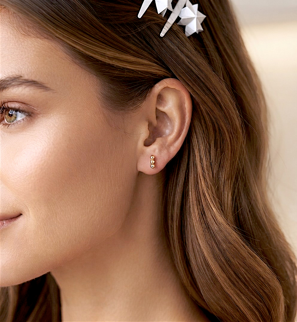 Close-up profile portrait of a woman with warm brown hair and a natural makeup look, smiling slightly. She is wearing a single 14K gold-filled triple bead vertical bar stud earring (as seen in image_5.png) in her earlobe. Her hair is pulled back to clearly display the minimalist, delicate gold cluster stud. The background is a soft, minimalist studio setting