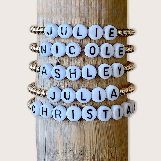 A collection of custom name bracelets for kids with gold beads and white bead letters, displayed on a wooden post.