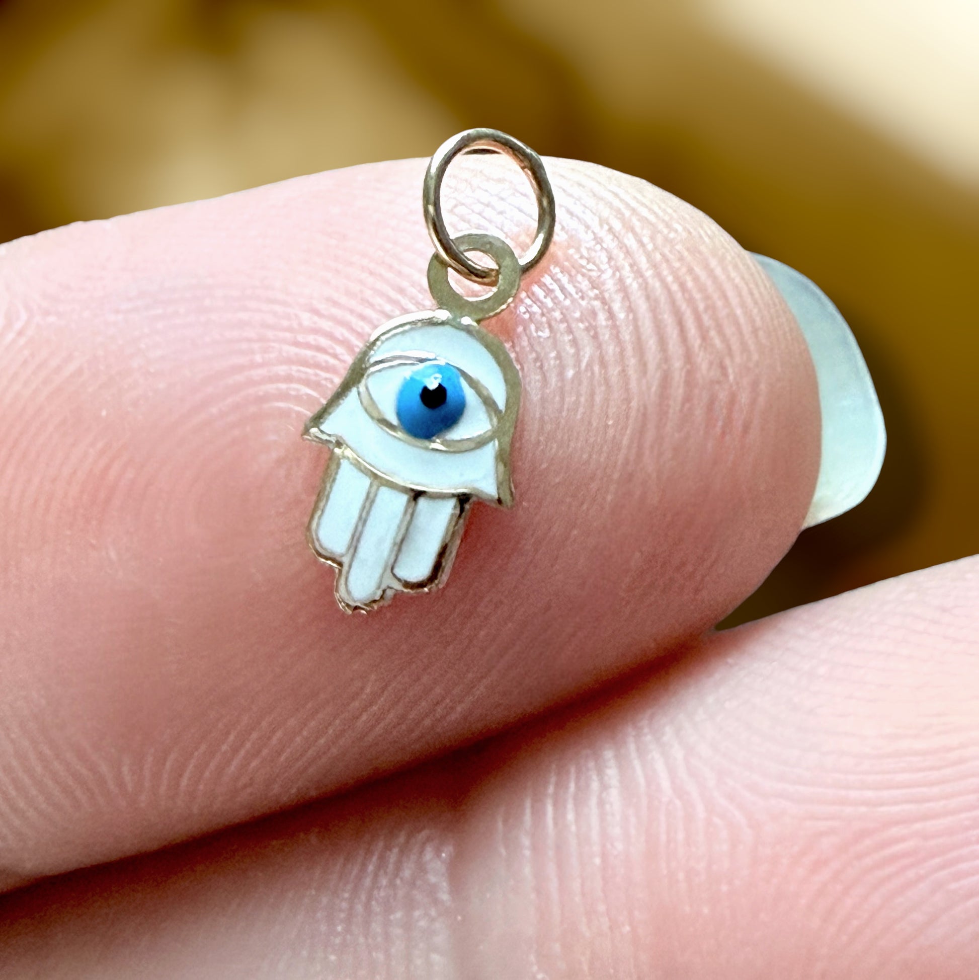 A 14K Gold Hamsa hand charm with a white enamel body and a blue-and-black evil eye detail, shown resting on a person's fingers.
