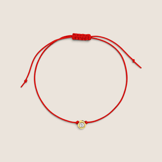 Red string bracelet with a small round gold bezel-set cubic zirconia, sliding knot closure, and two knotted cord ends on a warm neutral background