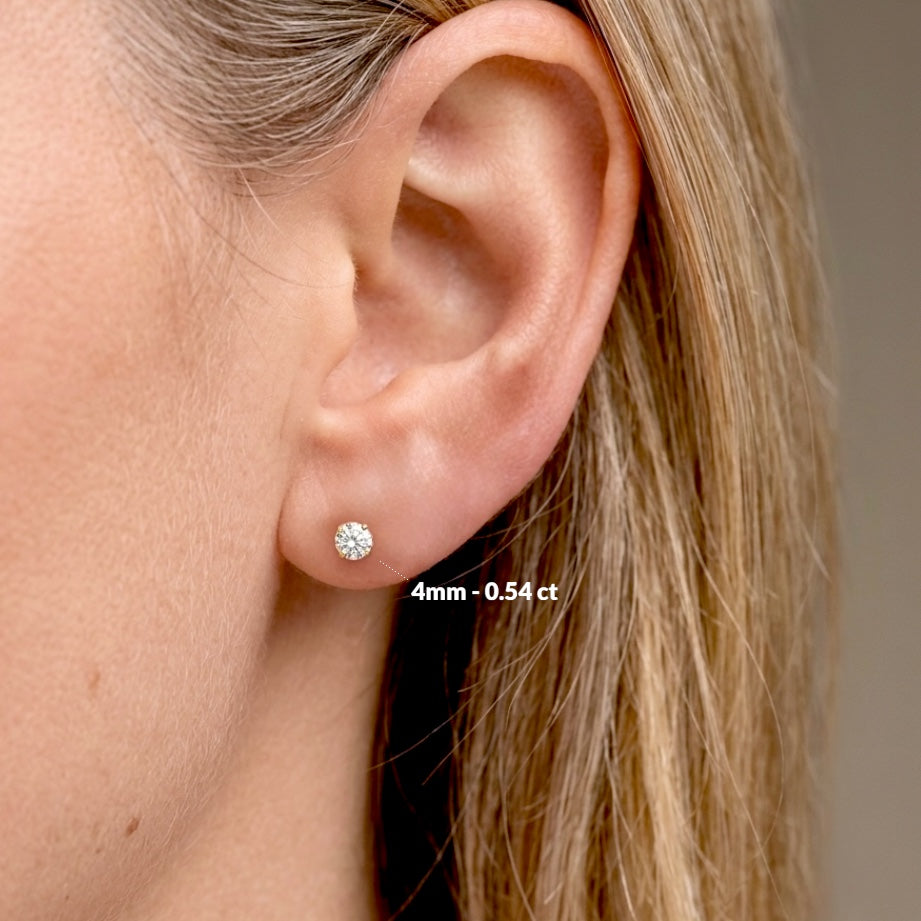 Close-up of an ear with diamond earrings and size label. Side view of real 14K solid gold round diamond studs in premium VS-VVS diamond clarity.Model wearing earrings for size visual - (4mm), 0.54 ctw
