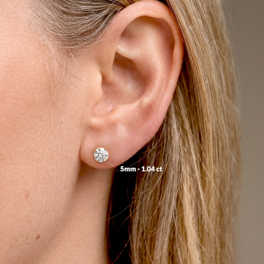 Close-up of an ear with diamond earrings and size label. Side view of real 14K solid gold round diamond studs in premium VS-VVS diamond clarity.Model wearing earrings for size visual - 1.04 ctw (5mm)