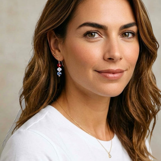 A close-up lifestyle photo of a model wearing a handcrafted 14k gold-filled patriotic drop earring featuring a vertically stacked red white and blue Swarovski crystal bead design on a French wire hook by Ashley Nicole Jewelry.