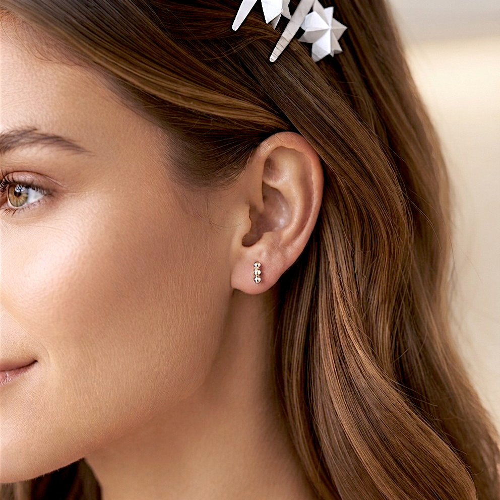 Close-up profile portrait of a woman with warm brown hair and a natural makeup look, smiling slightly. She is wearing a single Sterling Silver triple bead vertical bar stud earring in her earlobe. Her hair is pulled back to clearly display the minimalist, delicate polished cluster stud. 