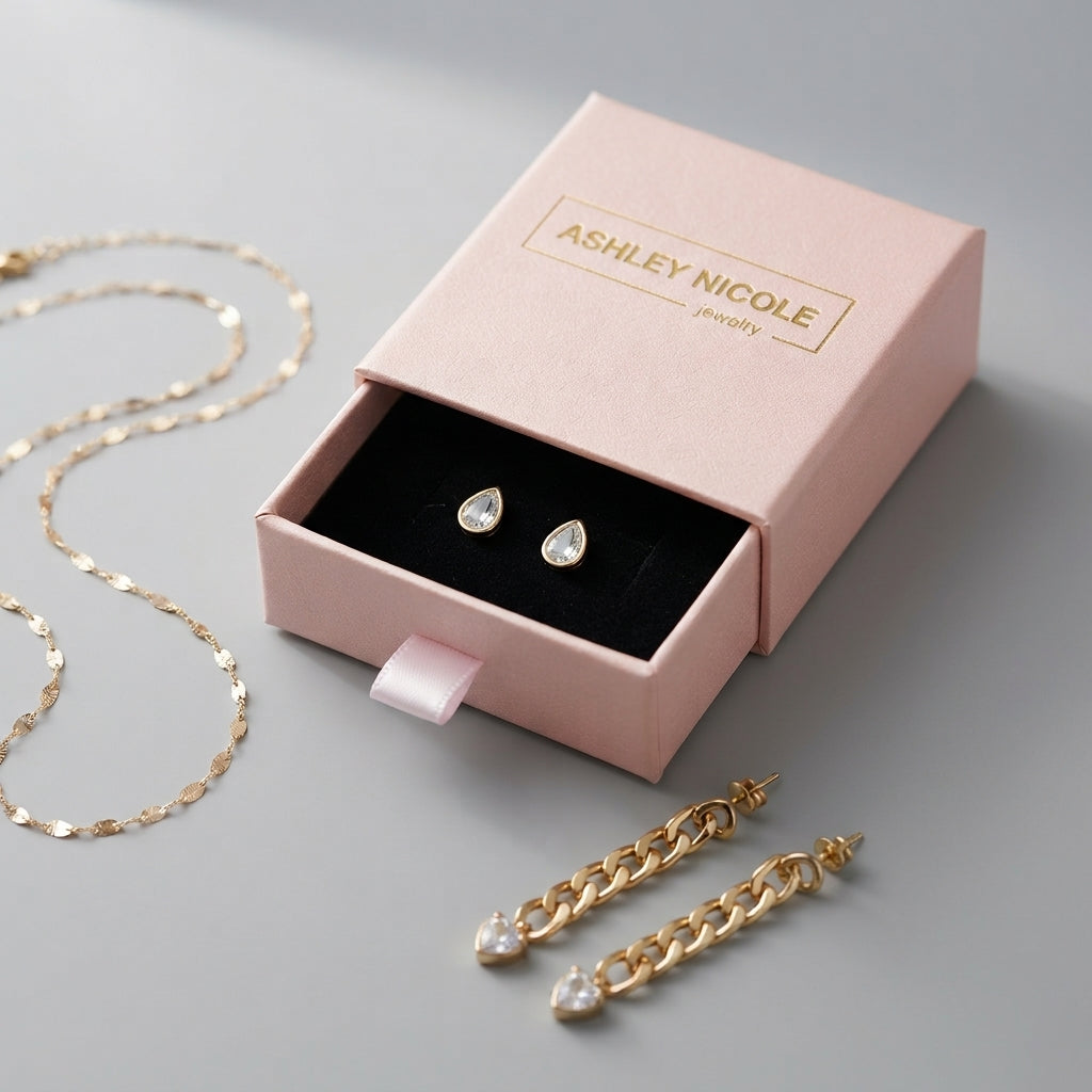 Open pink Ashley Nicole Jewelry gift box holding teardrop earrings, with a necklace and gold-tone drop earrings arranged around it.