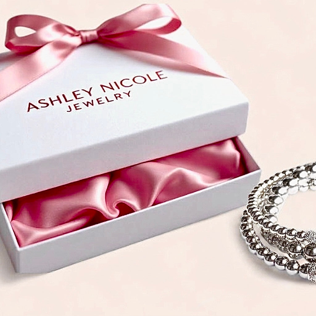 A crisp luxury white Ashley Nicole Jewelry gift box lined with a premium pink satin interior pouch, resting next to a stacked sterling silver bead bracelet.