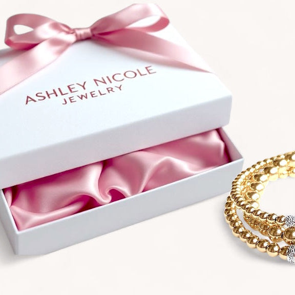 Gold bracelets on a white surface with an open jewelry box featuring a pink ribbon and 'Ashley Nicole Jewelry' branding.