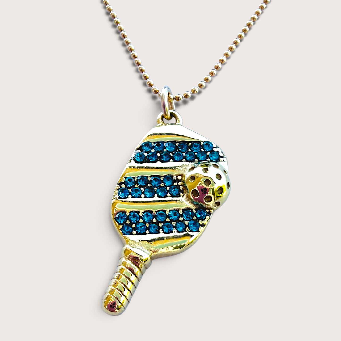 Gold pickleball paddle necklace with blue crystal accents displayed on a neutral background, highlighting the 1.0mm beaded chain.