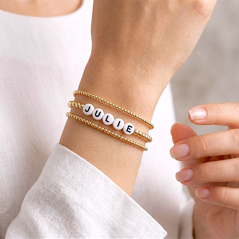 A close-up of a person's wrist wearing three stacked gold bracelets: two gold beaded bands and one gold band with white letter beads spelling 'JULIE'. By Ashley Nicole Jewelry 