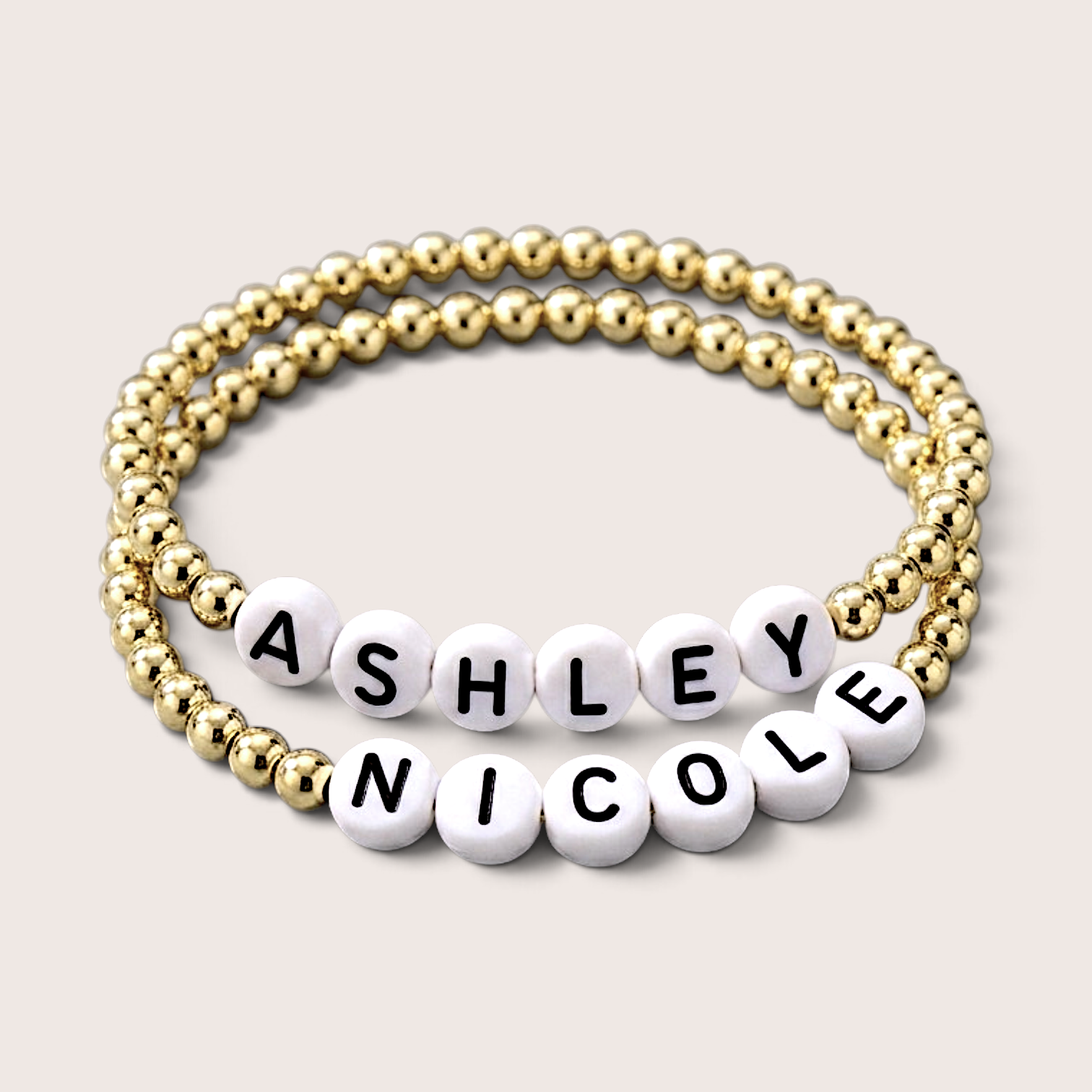 Custom 14K gold-filled  stretch beaded name bracelet with white letter beads. One bracelet is made of gold round beads, and the other features white round letter beads spelling out the names 'ASHLEY' and 'NICOLE' with black lettering.