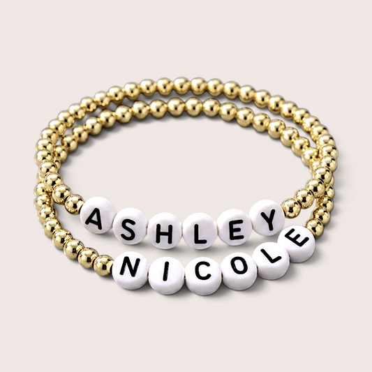 Custom 14K gold-filled  stretch beaded name bracelet with white letter beads. One bracelet is made of gold round beads, and the other features white round letter beads spelling out the names 'ASHLEY' and 'NICOLE' with black lettering.