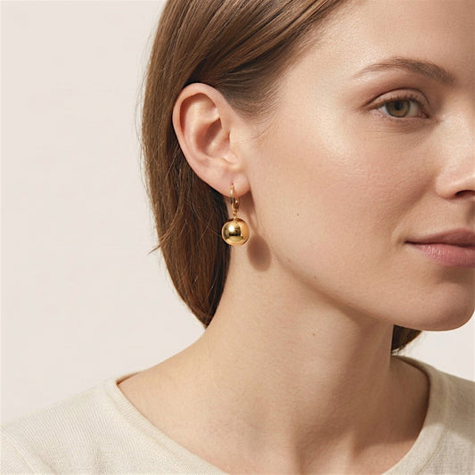 Woman with chestnut hair tucked behind her ear wearing a small polished gold ball drop earring and an ivory top.