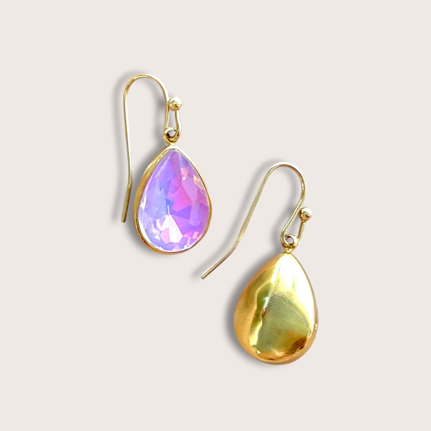 A pair of gold teardrop dangle earrings, one with a faceted iridescent purple gemstone and the other with a smooth gold gemstone, set against a light beige background.
