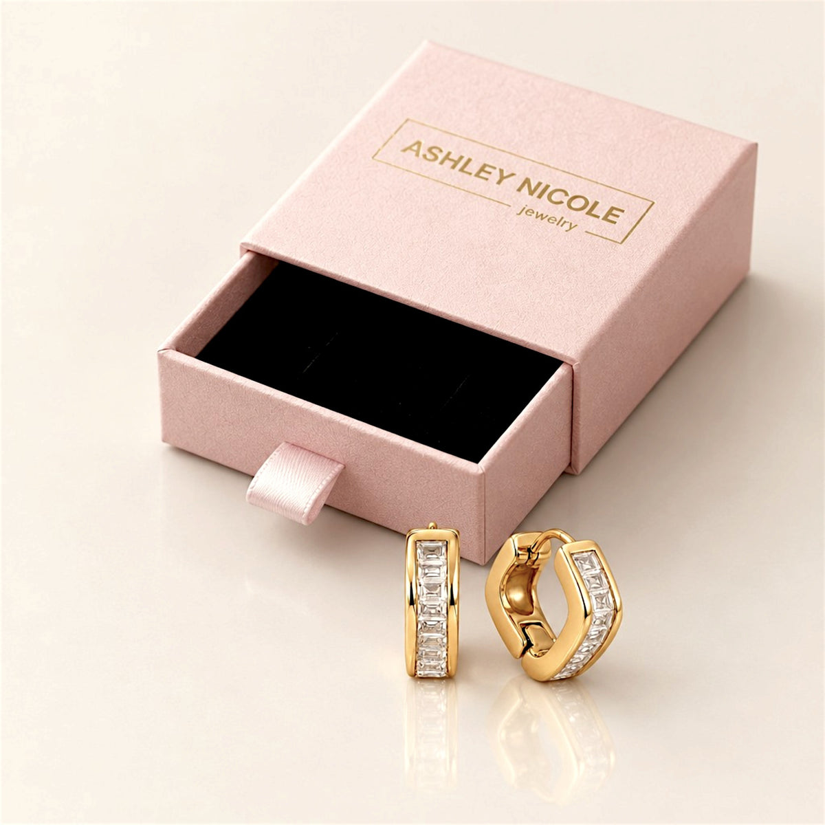 Gold hex baguette huggie hoop pair beside an open pink Ashley Nicole Jewelry drawer-style gift box.