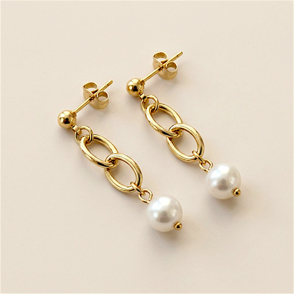 Two gold link pearl drop earrings arranged diagonally on a neutral surface, with the posts and butterfly backs visible.