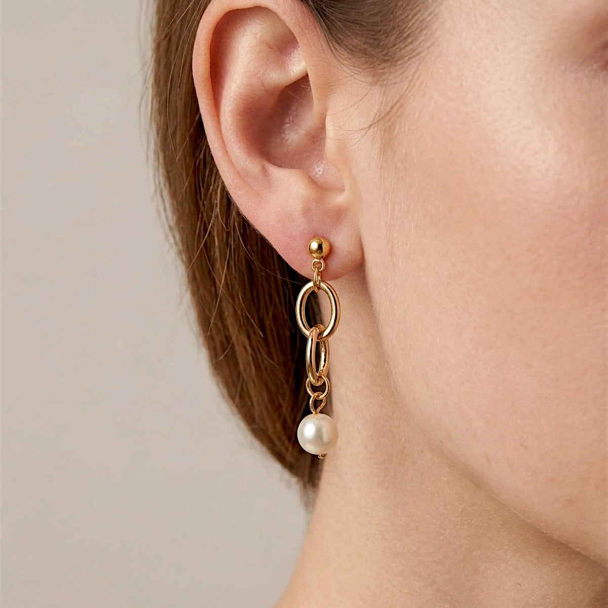 Close-up of a gold pearl drop earring worn on the ear, showing the ball stud, interlocking oval links, and small pearl connectors.