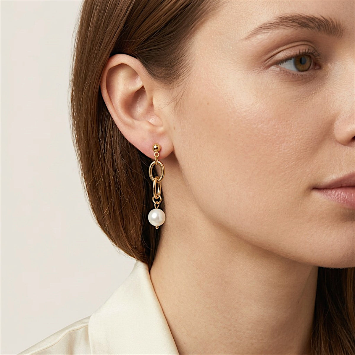 Brown-haired woman wearing a gold link pearl drop earring, with her hair tucked behind her ear and an ivory collared shirt.