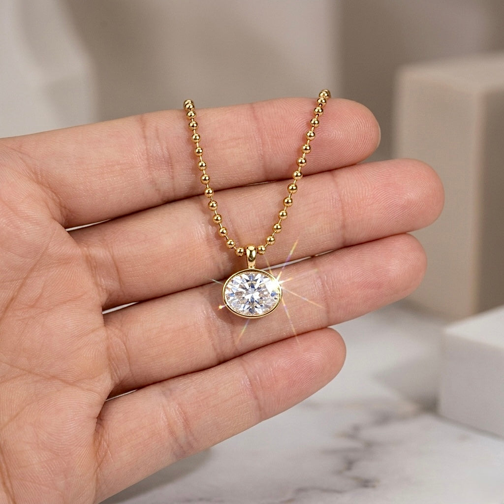 Female hand holding the 18K gold east-west oval bezel CZ pendant necklace, showing the scale and sparkle in natural light against a blurred white marble background.