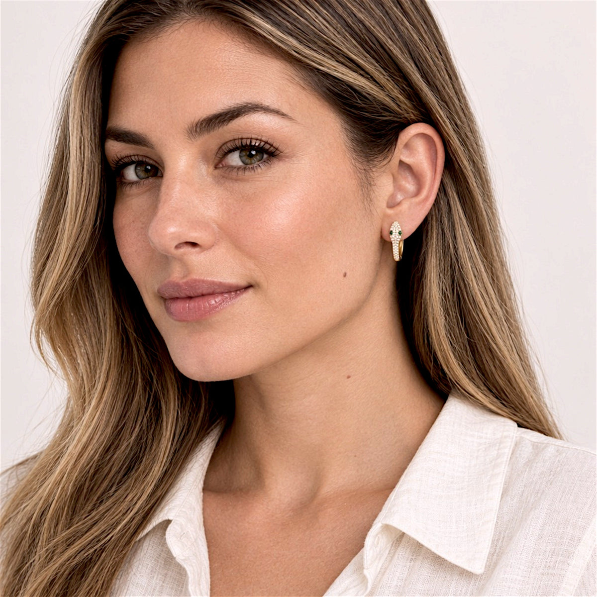 Lighter-haired model in an ivory blouse wearing a gold pavé snake huggie hoop, with her hair tucked behind her ear.