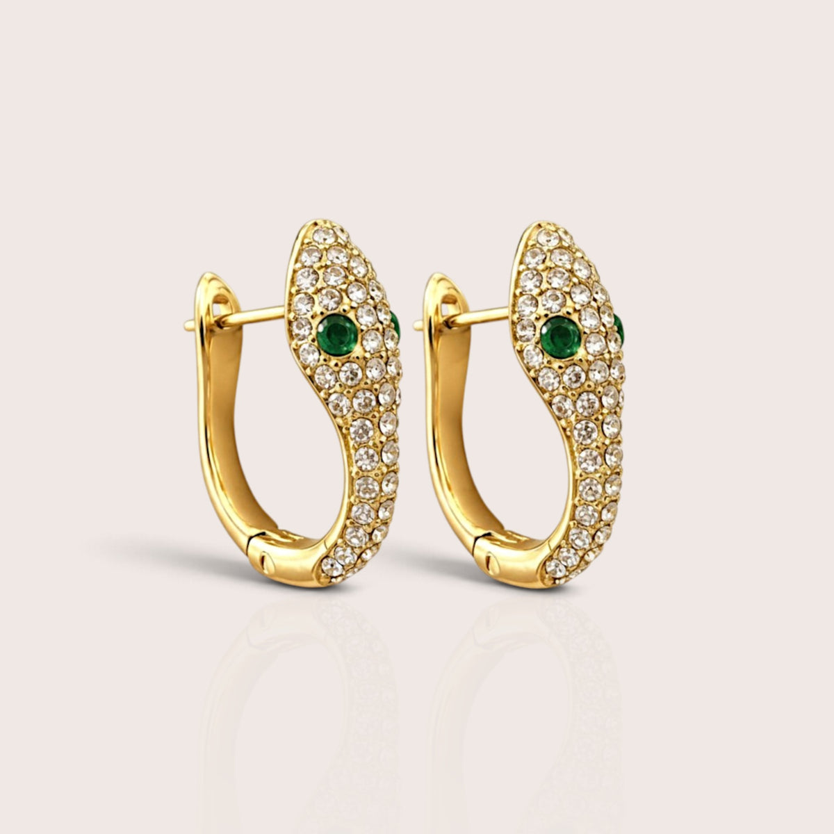 Pair of gold U-shaped snake huggie hoops with clear pavé zircon stones and green zircon eyes on a neutral background.