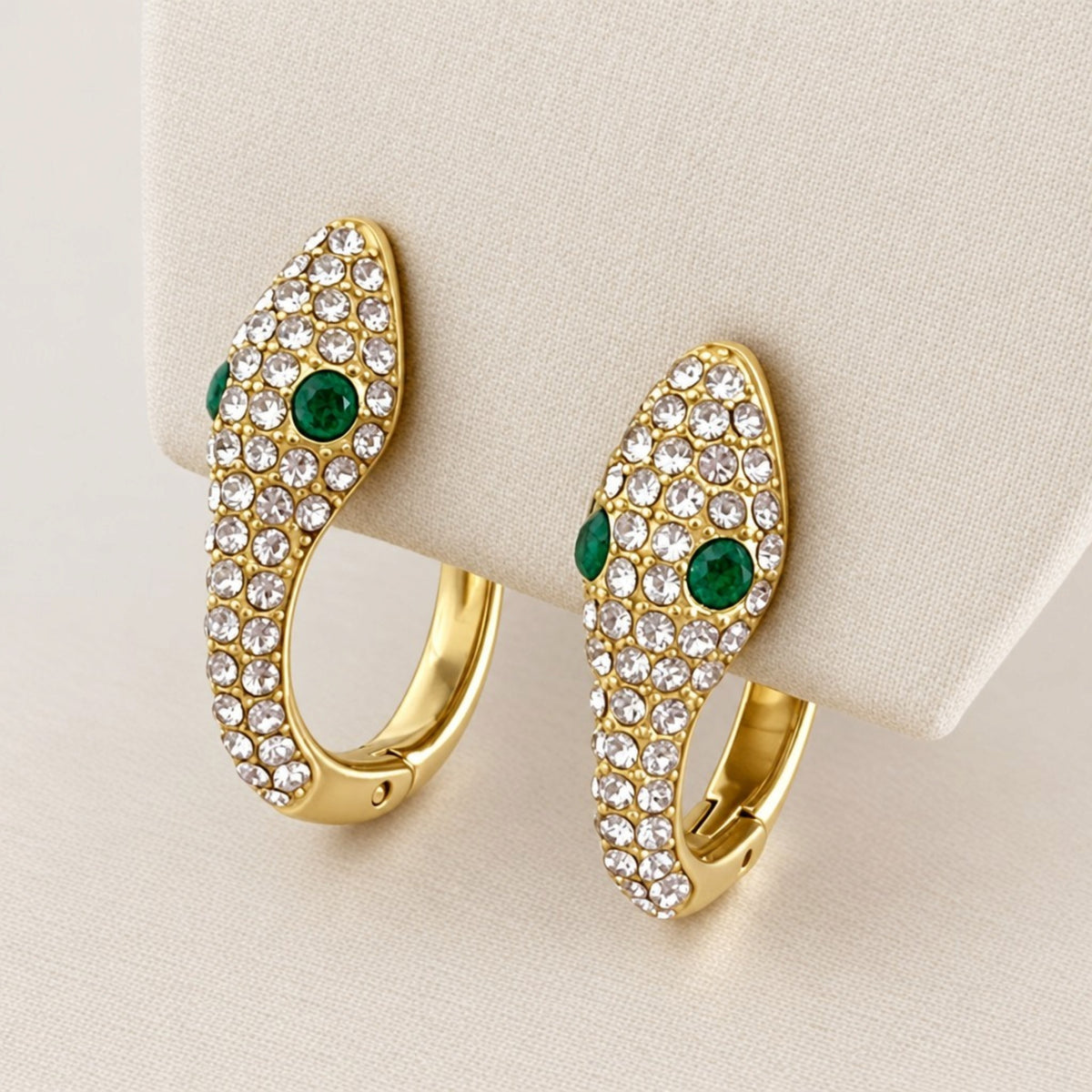 Gold snake huggie hoop pair on a cream fabric display, showing clear pavé zircon stones, green eyes, and polished gold backs.