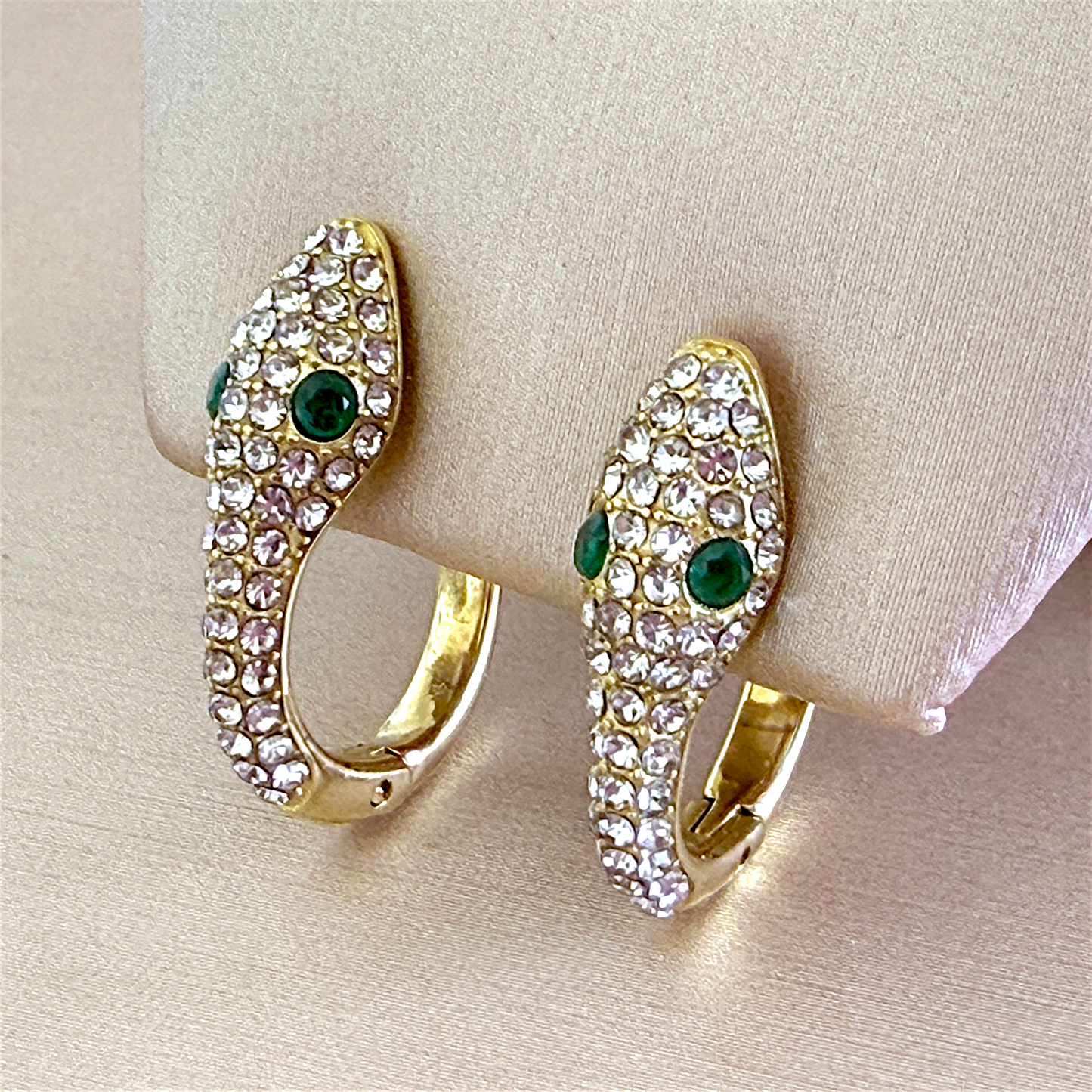 A pair of 18K gold PVD-finished pavé snake huggie earrings mounted on a cream-colored jewelry display stand, highlighting the symmetry and encrusted clear zircons.