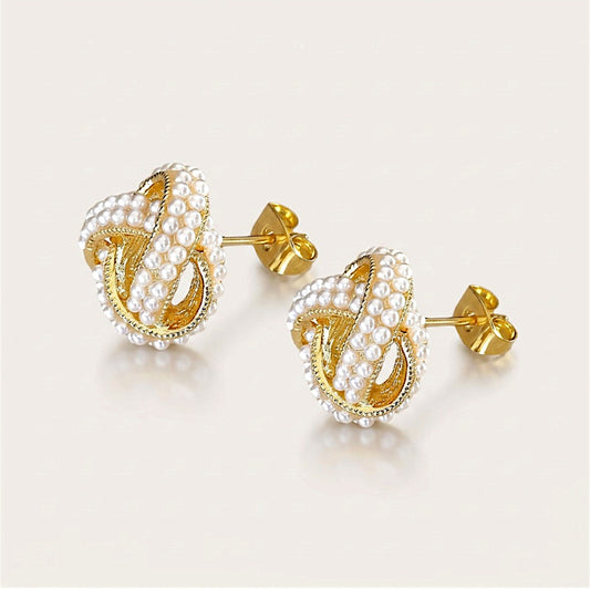 Pair of gold pearl love knot stud earrings shown at an angle, with textured gold edging, straight posts, and butterfly backs visible.