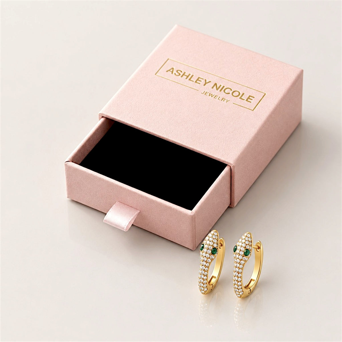 Gold pavé snake huggie hoop pair beside an open pink Ashley Nicole Jewelry drawer-style gift box with a black insert.