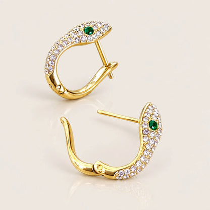 Gold snake huggie hoops at two angles, showing clear pavé zircon, green eyes, U-shaped profiles, and an open hinged closure.