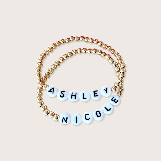 Gold beaded bracelets with name 'ASHLEY NICOLE' on a beige background