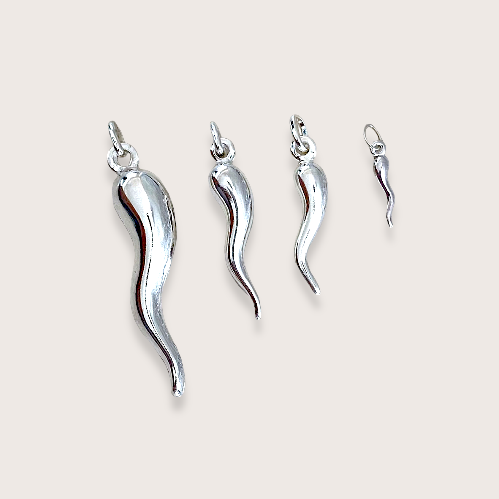 Four sizes of the solid 925 sterling silver cornicello Italian horn pendant charms by Ashley Nicole Jewelry arranged in a neat row from large to mini on a soft cream background.