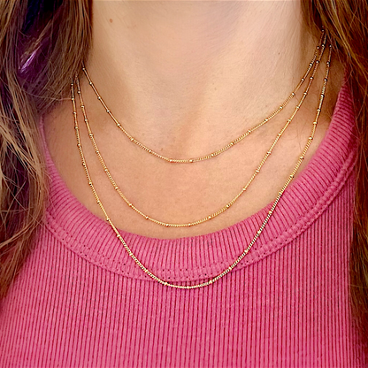 Lifestyle portrait of a model wearing multiple layered 14K gold-filled beaded satellite station necklaces styled together against a pink textured top
