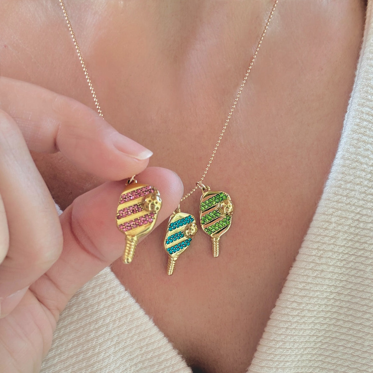 Hand holding the gold pickleball necklace with three multi-colored paddle charms against a neutral background.