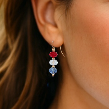 A close-up lifestyle photo of a model wearing a handcrafted 14k gold-filled patriotic drop earring featuring a vertically stacked red white and blue Swarovski crystal bead design on a French wire hook by Ashley Nicole Jewelry.