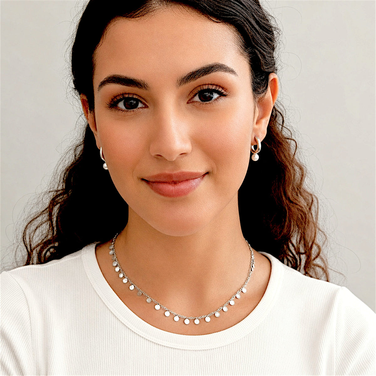 Model wearing silver pearl hoop earrings with a white top and a silver disc necklace.