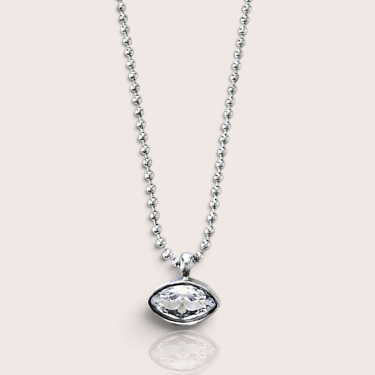A silver beaded necklace with an oval bezel-set clear gemstone pendant, shown against a solid light beige background.