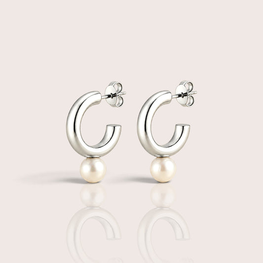 Pair of chunky silver open C hoop earrings with white pearl drops, shown on an ivory surface with soft shadows and reflections.