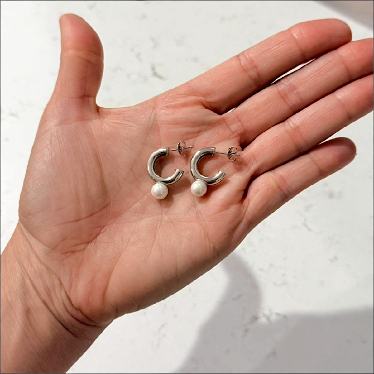 Pair of silver pearl C hoop earrings resting in an open palm, showing their size relative to the hand.