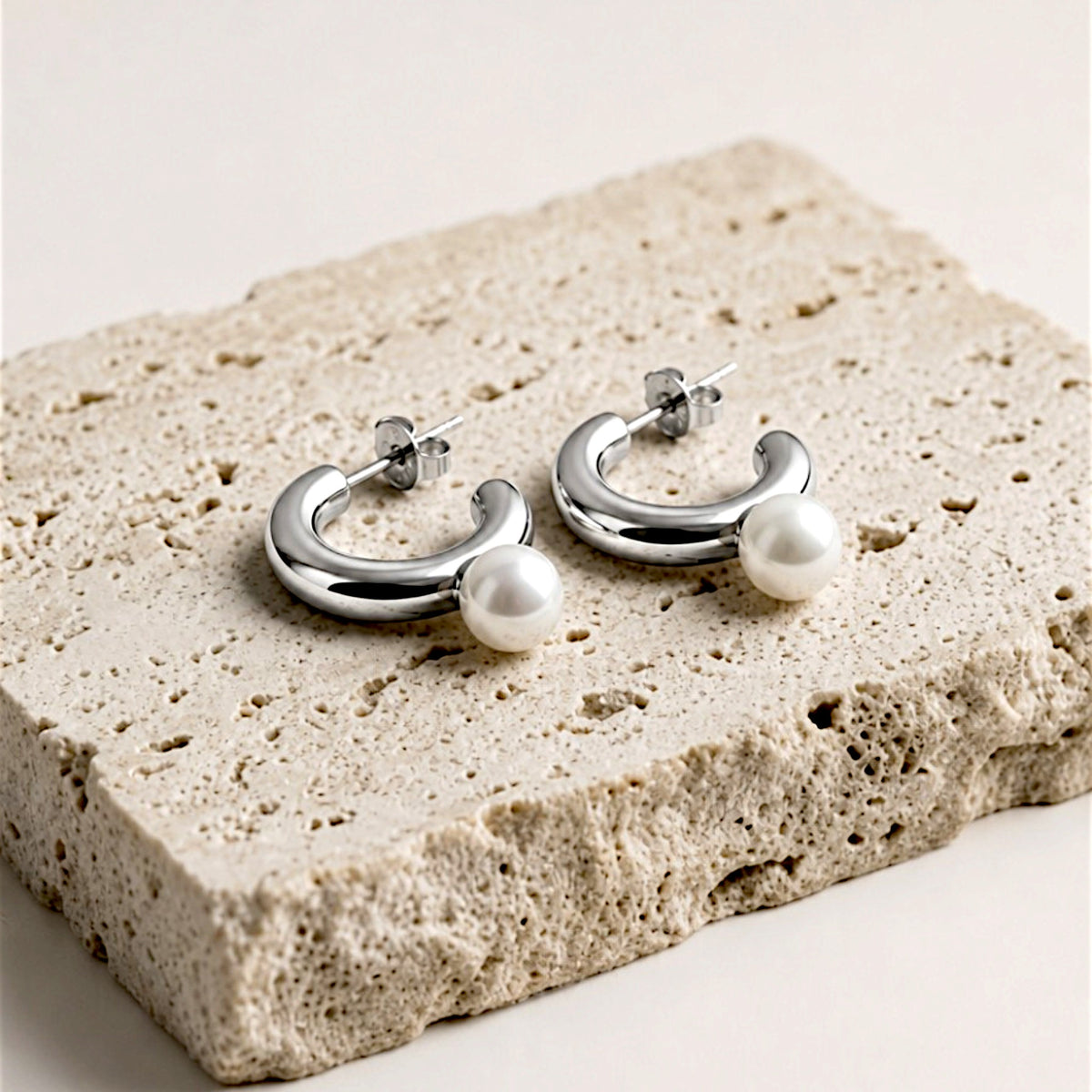 Pair of silver pearl C hoop earrings resting on a textured beige stone slab, with posts and butterfly backs visible.